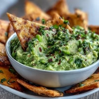 Avocado-Edamame-Dip mit knusprigen Pita-Chips in einer Schüssel, serviert mit goldbraunen Dreiecken daneben.